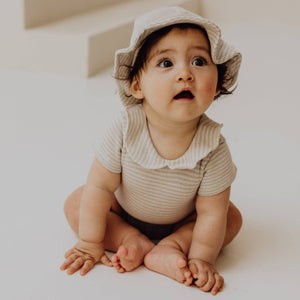 Baby wearing a striped body with collar and a sun hat, seated on a soft surface, showcasing a cute and stylish look.