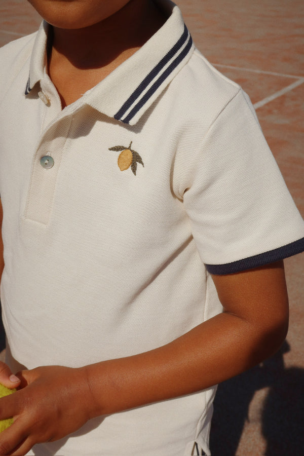 Child wearing Florian Poloshirt in Antique White with blue trim, featuring a lemon embroidery.