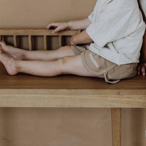 Beige Musselin-Shorts für Kinder auf einer Holzbank, leicht und bequem für warme Tage.