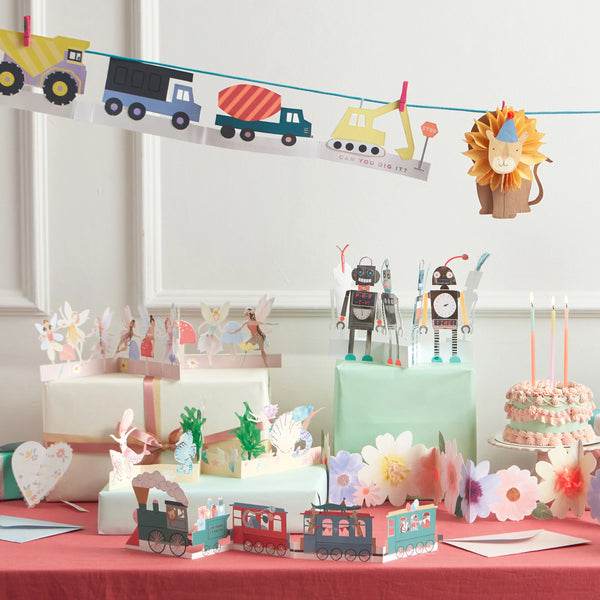 A colorful birthday party setup featuring a train-themed centerpiece, robot decorations, pastel-colored cakes, and gift boxes with floral accents.