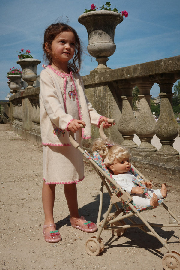 Child in a creamy cardigan and dress pushing a stroller with dolls in a sunny outdoor setting.