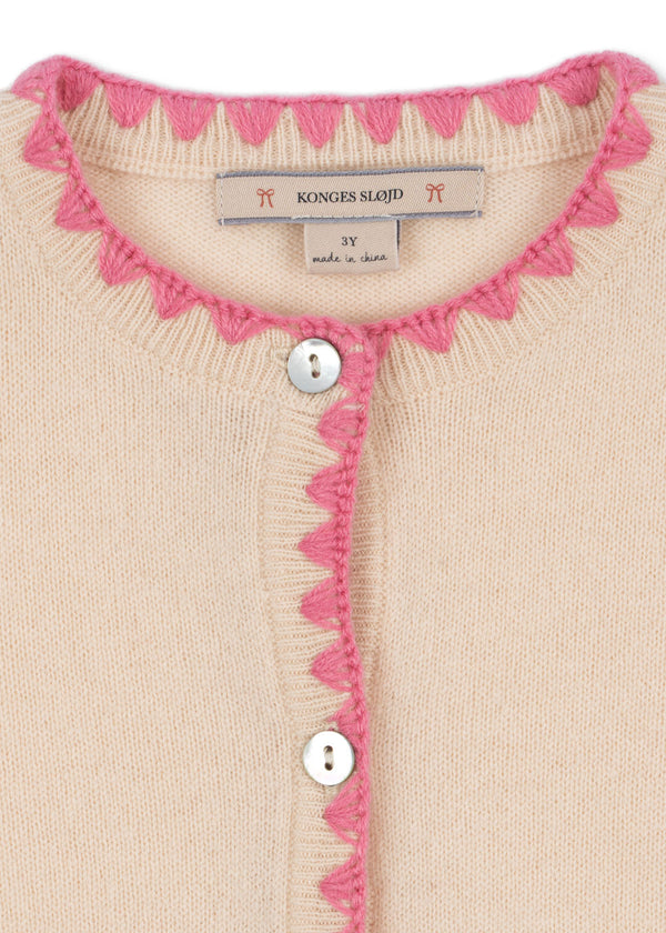 Close-up of Konges Sløjd off-white cardigan with pink scalloped trim and pearl buttons, showcasing delicate craftsmanship.